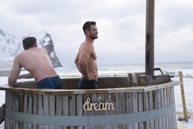 Two men in a wooden hot tub outdoors, overlooking the ocean.