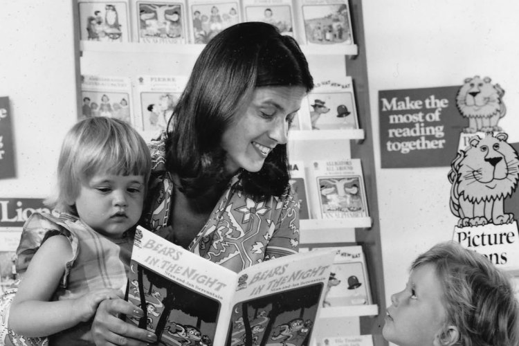 Black and white photo of a woman reading "Bears in the Night" to two young girls in front of a Picture Lions book display.