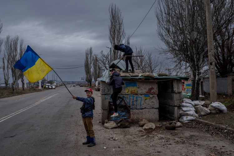 Ukrainian children playing at an abandoned checkpoint in Kherson, waving a Ukrainian flag.