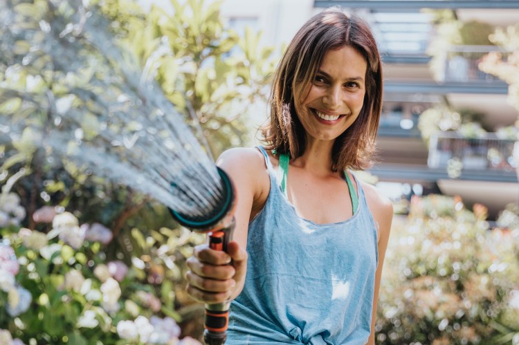 Woman smiling while watering plants with a garden hose.