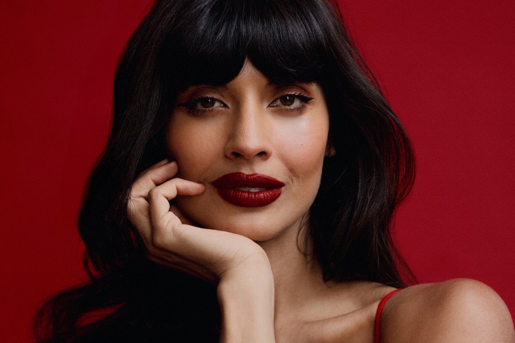 Jameela Jamil in a red dress, seated on a red background.