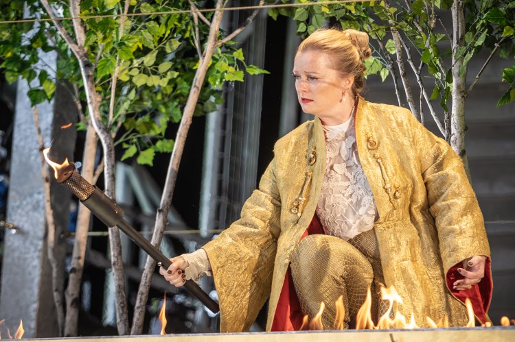 Lucy Crowe as Rodelinda in a gold-colored costume, holding a torch.