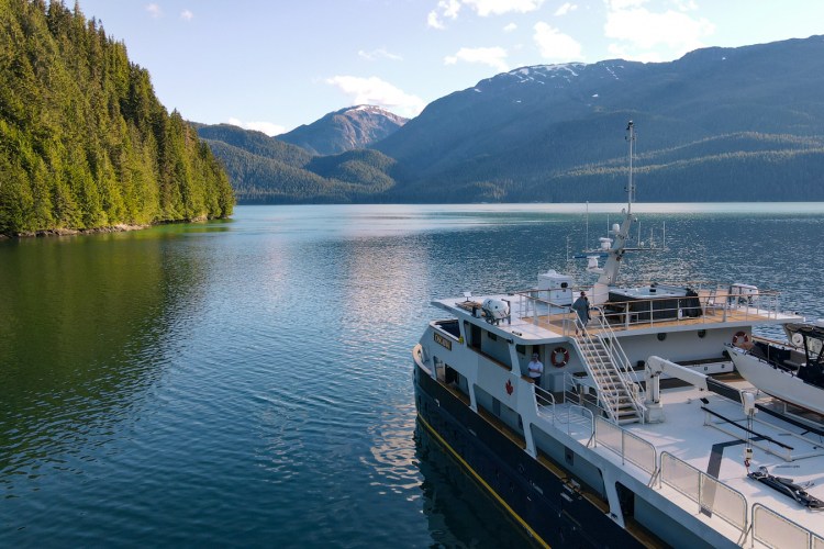 The Cascadia, a ship, on a calm body of water surrounded by mountains and lush green forests.